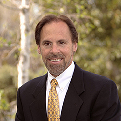 The image shows a man wearing a suit with a yellow tie, smiling, and posing for a portrait against a background with trees and a clear sky.