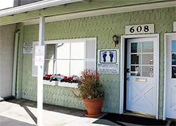 The image shows an exterior view of a building with a sign indicating a business or service, featuring a door with a number 608 above it, a potted plant on the porch, and a small sign in front of the entrance. The building has a green facade and is surrounded by a parking lot. There are no visible people or moving objects in the image.